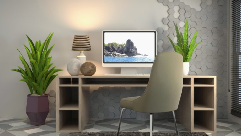 A modern, stylish workspace tailored for a dynamic workforce features a wooden desk with an Apple iMac displaying a seascape. Two potted plants are on the desk, and two more large plants flank the desk. A gray chair is positioned in front of the desk. Hexagonal wall tiles provide a unique backdrop.