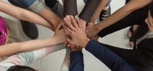 A diverse group of people standing in a circle, each extending one hand into the center to join with the hands of others, symbolizing unity and teamwork. The photo beautifully captures diversity through the various skin tones, clothing styles, and genders of the participants.