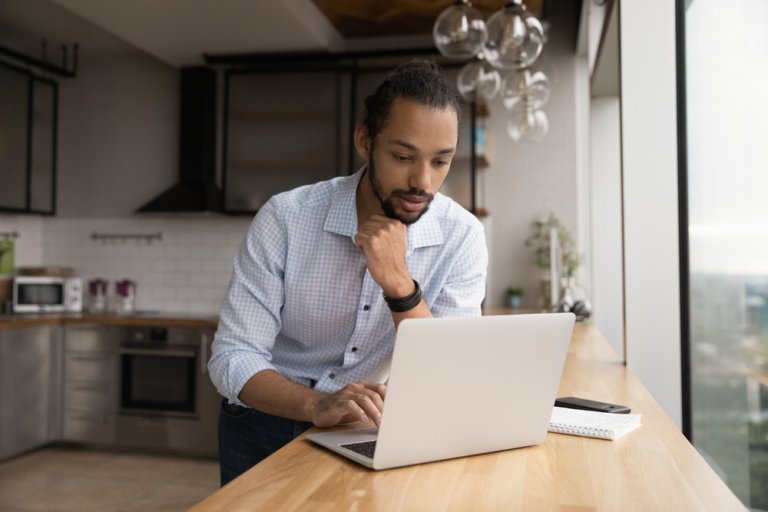 A young man standing by a desk and looking at a laptop