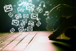 A close-up of a person's hand using a laptop's trackpad, with glowing envelope icons floating above the keyboard, symbolizing emails. The background is dark, and other icons like music notes and gears are faintly visible, representing different applications, including government services.