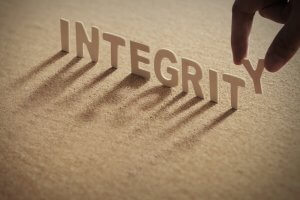 A close-up of the word "INTEGRITY" in white 3D letters on a textured surface. A hand is adding the letter "Y" at the end, casting elongated shadows from the letters onto the surface due to the lighting.