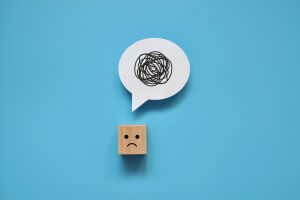 A wooden block with a sad face drawn on it is positioned at the bottom center of a blue background. Above it, a white speech bubble contains a scribbled, tangled line, symbolizing confusion or frustration often caused by difficult people.