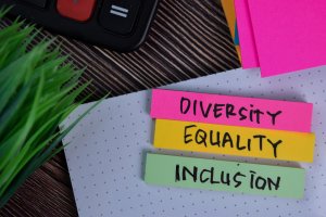 A close-up of sticky notes arranged on dotted paper, with each note reading "DIVERSITY," "EQUALITY," and "INCLUSION." In the background, there is a potted plant and part of a calculator visible. The scene subtly underscores the importance of diversity in any setting.