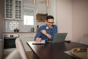 A man wearing glasses and headphones is sitting at a dining table with a laptop, notepad, and pen. Smiling, he appears to be engaged in an online meeting or video call related to government affairs in a modern kitchen setting.