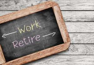 A small chalkboard with a wooden frame displays the words "Work" and "Retire" written in chalk, with arrows pointing in opposite directions. The chalkboard rests on a weathered wooden surface, reminiscent of an old government office desk.