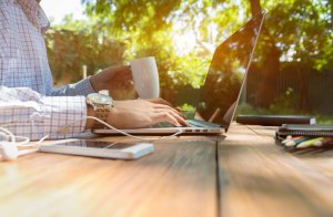 A person engaged in remote working sits at a wooden table outdoors, holding a white mug in one hand. Sunlight filters through the trees, creating a serene, productive setting. A smartphone, notebook, and pen lie on the table beside their laptop.