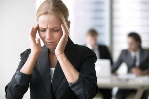 A woman in a black blazer stands holding her temples, appearing stressed or experiencing a headache. In the background, two blurred colleagues in business attire work at a desk in an office setting, possibly indicating the presence of difficult people.