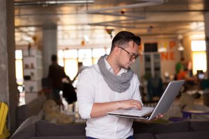 A man with short hair, wearing glasses, a white shirt, and a grey scarf is standing indoors and working on a laptop. He is focused on the screen, amidst the bustling workforce of a modern, open-plan office space in the background, which includes a sofa and various office furniture.