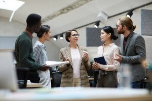 A diverse group of five professionals are standing and having a discussion in a modern office setting. They are dressed in business casual attire, and one person is holding a clipboard while another has a notebook. The office, reflecting their diversity, has bright lighting and contemporary decor.
