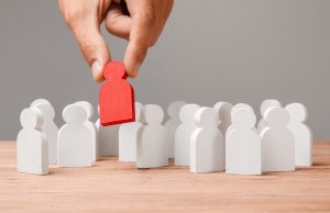 A person's hand is lifting a red figure from a group of white figures on a wooden surface, symbolizing selection or leadership among many, much like government recruitment. The background is plain gray.