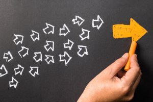 A hand is drawing a large, solid yellow arrow with chalk on a blackboard, pointing to the right. Numerous smaller, outlined white arrows point towards the yellow arrow, symbolizing leadership and creating a sense of movement and direction toward the right side of the image.