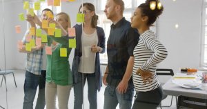 A group of five people are collaborating, standing and looking at sticky notes on a glass wall in an office setting. Some of them are pointing at the notes, while another person holds a cup. The room is filled with bright, natural light, enhancing the focused and engaged atmosphere.