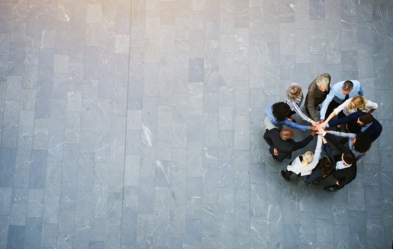 An aerial view captures a diverse group of people standing in a circle on a gray tiled floor, reaching into the center to stack their hands together, symbolizing unity, collaboration, and teamwork. The photo is taken from above, showing a significant amount of empty space around the group.
