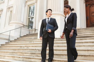 A man and a woman, both dressed in business attire, are walking down the steps of a large government building with tall columns. They appear to be engaged in conversation and smiling. The man holds a folder, suggesting they might be colleagues or professionals.