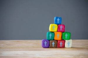 A pyramid of playdough blocks in various colors sits on a wooden surface against a plain gray background. Each block features a small white stick figure icon, symbolizing teamwork. The pyramid, ideal for team building activities, consists of ten playdough blocks in total.