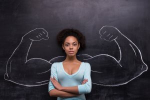 A confident woman with arms crossed in front of a chalkboard drawing of large, muscular arms flexed on either side of her. She wears a light blue top and stands confidently, exuding leadership and giving the impression of inner strength and empowerment.