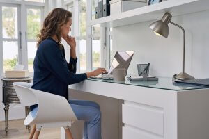 A woman with long hair is sitting at a white desk in a modern home office, engaged in remote work. She is using a laptop and appears to be concentrating. The desk has various office supplies, and there is a lamp on it. Shelves with books and binders are in the background.