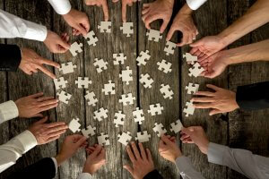 A group of people, primarily showing their hands, collaborate on assembling scattered white puzzle pieces on a wooden table. Hands are reaching in from all sides, indicating teamwork and cooperation in this team-building activity to solve the puzzle.