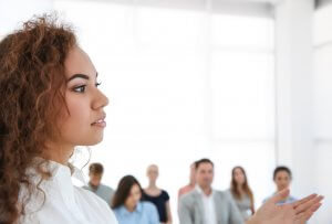A woman with curly hair, dressed in a white shirt, stands and speaks in front of a group of people. The audience, slightly blurred in the background, listens attentively. The setting appears to be a modern, bright, and well-lit room or office, reflecting her leadership role.
