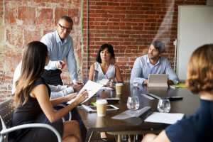 A group of five people is having a collaborative meeting in a modern office with exposed brick walls. One person is standing and appears to be leading the discussion, while the others are seated around the table, engaged and taking notes.