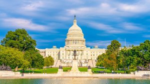 The United States Capitol building in Washington, D.C., is bathed in sunlight. The dome is prominently featured, with the National Mall and reflecting pool in the foreground, surrounded by green lawns and trees. The sky is partly cloudy—a reminder of bustling landscapes from government jobs in PA.