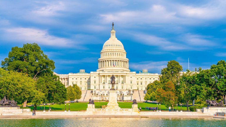 The United States Capitol building in Washington, D.C., is bathed in sunlight. The dome is prominently featured, with the National Mall and reflecting pool in the foreground, surrounded by green lawns and trees. The sky is partly cloudy—a reminder of bustling landscapes from government jobs in PA.