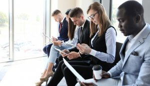 A group of professionally dressed people sit in a row in an office waiting area. They are focused on documents and electronic devices, with one person holding a coffee cup. Large windows in the background let in natural light, providing a view of the outdoors. They are likely candidates for jobs in the government.