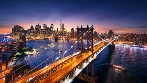 A breathtaking view of Manhattan, New York City, at dusk. The skyline is illuminated with lights, featuring the iconic One World Trade Center. The Brooklyn Bridge and Manhattan Bridge are visible, with a river reflecting the vibrant city lights—a stark contrast to wondering how many jobs are available in military/government/technical fields.