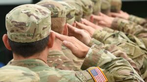 A group of soldiers in camouflage uniforms, viewed from the back, standing in line and saluting. One soldier's sleeve displays a U.S. flag patch, symbolizing dedication to their mission and representing some of the best paying jobs in military/government/technical sectors.