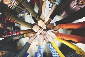A diverse group of people standing in a circle reach their hands towards the center, connecting them in a gesture of unity and teamwork. The image is taken from above, showcasing various clothing styles and skin tones.