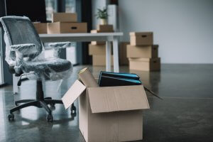 An empty office with a brown cardboard box containing binders and a rolled-up poster in the foreground. A black office chair covered with plastic wrap is near a desk in the background. Stacked cardboard boxes, labeled for government use, are visible against the walls.