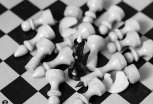 A black chess king standing upright amidst a scattered pile of white chess pieces on a chessboard. The fallen white pieces surround the black king, emphasizing its dominance and leadership. The image is in black and white.