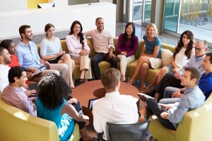 A diverse group of people, seated in a circle in a modern, bright office space, engage in a discussion. They appear relaxed and attentive, suggesting a collaborative meeting or team-building session that highlights the positive work culture. A reception desk and large windows are visible in the background.