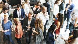 A diverse group of people mingling at a social event, connecting over lively conversations while holding drinks. Others engage in small group discussions. The setting appears to be a modern indoor venue with a welcoming atmosphere.