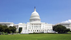 The image shows the United States Capitol building in Washington, D.C., set against a clear blue sky. It is a grand white structure with a large dome in the center, flanked by long wings on either side, and a well-maintained lawn in the foreground—an apt setting for discussing the pros and cons of government jobs in the USA.