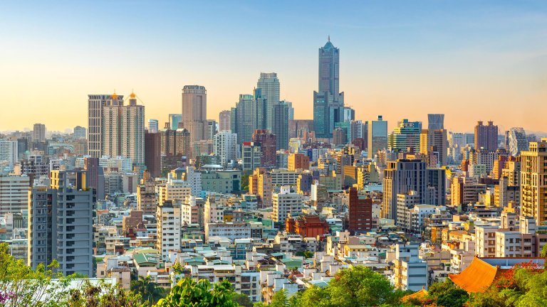 A cityscape of a modern urban area during sunset, featuring a dense cluster of high-rise buildings and skyscrapers. The tallest skyscraper stands prominently in the center. The sky is clear with a warm gradient from blue to orange, casting a gentle glow over the city.