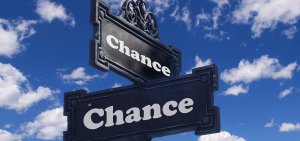A street signpost against a bright blue sky with fluffy white clouds displays two black signs with ornate borders, both reading "Chance" in bold white letters, one pointing left and the other pointing right. This imagery evokes the pros and cons of government jobs in the USA, each direction offering its own path.