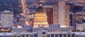A view of a cityscape at dusk with the illuminated dome of the Utah State Capitol building prominently in the center. The building is surrounded by various tall buildings, and a long stretch of street lights can be seen extending into the distance. Ready to apply to government jobs in NC? Don't miss out!