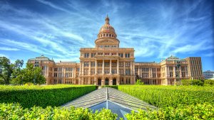 A grand, sandstone-colored capitol building stands under a vibrant blue sky with wispy clouds. The structure features a prominent central dome and symmetrical wings. Searching for government jobs in Texas for something special? Manicured hedges and a geometric pathway lead toward the majestic building.