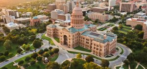 Aerial view of the Texas State Capitol building in Austin, surrounded by lush green lawns and trees. The grand, historical structure, perfect for those searching government jobs in Texas for something special, is set against a backdrop of city buildings under a clear sky, with roads and pathways encircling the area.
