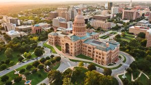 Aerial view of the Texas State Capitol building surrounded by lush greenery and trees. The historic dome and architecture are prominent, set within an urban downtown area with various buildings and skyscrapers in the background under a warm, late afternoon light. Searching government jobs in Texas for something special?