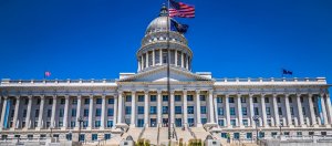 A grand neoclassical government building with a large dome, the Utah State Capitol, stands under a clear blue sky. The U.S. flag and Utah state flag wave atop flagpoles in front of the building. The facade features tall columns and a broad staircase leading up. Ready to apply to government jobs in NC? Don't miss out!