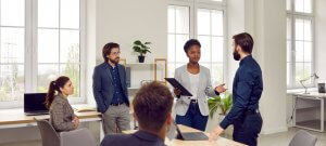 A group of five office workers are engaged in a discussion in a modern, well-lit office space, highlighting their collaborative work culture. One woman holding a tablet is speaking to a man in a dark shirt, while three other colleagues listen attentively. Large windows and office furniture are visible in the background.