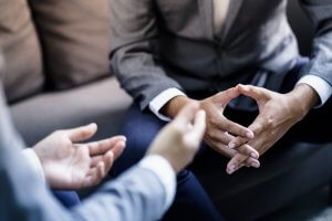 Two people in business attire are seated on a couch, engaging in an employment discussion. One person is gesturing with their hands, while the other has their fingers clasped. The focus is on their hands and body language rather than their faces.