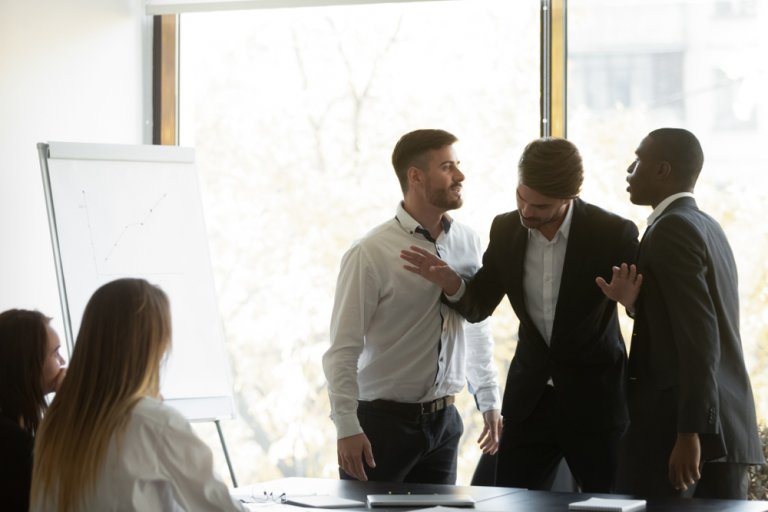 Two men are having a heated argument in an office meeting while a third man tries to mediate, attempting to prevent any escalation into workplace violence. Three other people, two women and one man, are seated and looking on. A whiteboard with a graph is visible in the background, and natural light streams through a large window.