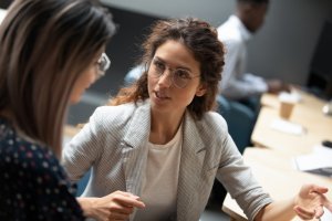 Two women are engaged in a conversation about employment in an office setting. One wears a gray blazer and glasses with curly hair sitting on the right, speaking intently. The other, with long, straight hair and glasses, is partially visible on the left. Desks are in the background.