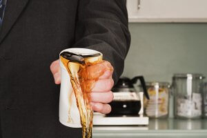 A person in a suit is pouring coffee from a pot into a white, disposable cup, seemingly spilling it over the side. Amidst the calm kitchen counter setting with a coffee maker and various containers, there's an underlying tension hinting at the stress of workplace violence.