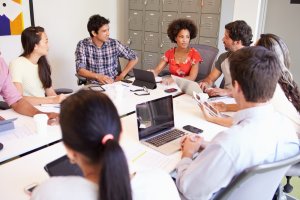 A group of people are gathered around a conference table in an office setting, reflecting a vibrant work culture. They are engaged in discussion, with some using laptops, tablets, and notebooks. Lockers are visible in the background, and one person in a red patterned shirt is speaking.