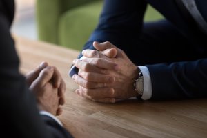 Close-up of two individuals sitting across a wooden table, each with hands clasped. Both are dressed in dark suits, and one wears a wristwatch. The image conveys a serious or formal discussion, possibly during a government meeting or interview.
