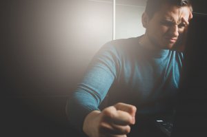 A man appears frustrated and stressed, holding his forehead with one hand and clenching his other into a fist. He is sitting in front of a laptop in a dimly lit environment, the tension hinting at underlying concerns of workplace violence.